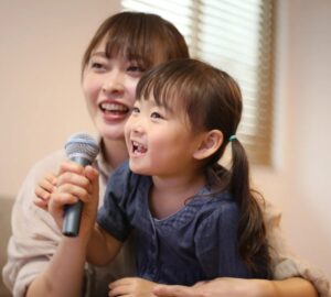 マイクを手にした幼い子どもが、そばで見守る大人と一緒に楽しそうに歌っている様子。