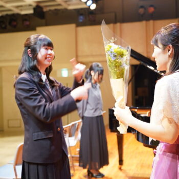 ピアノ発表会の舞台で、講師から生徒たちへ花束が手渡され、あたたかな交流が生まれている場面。
