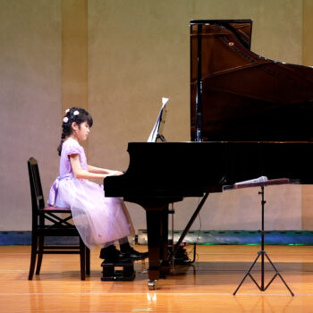発表会のステージで、ドレスを着た小さな生徒がグランドピアノを一生懸命演奏している様子。